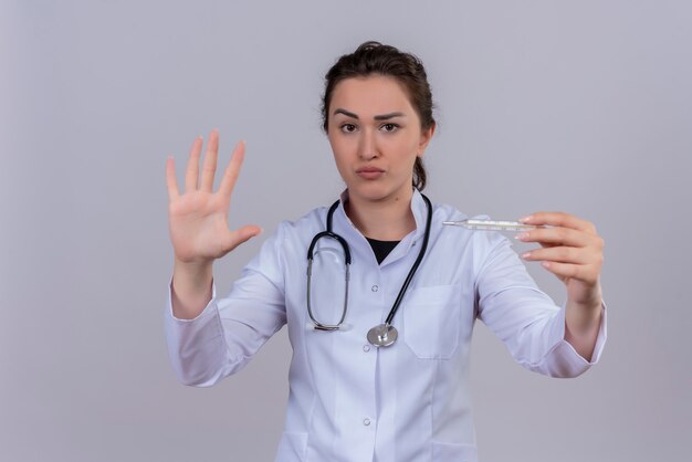 Regardant la caméra jeune médecin portant une robe médicale portant un stéthoscope tenant un thermomètre et montre un geste d'arrêt sur un mur blanc
