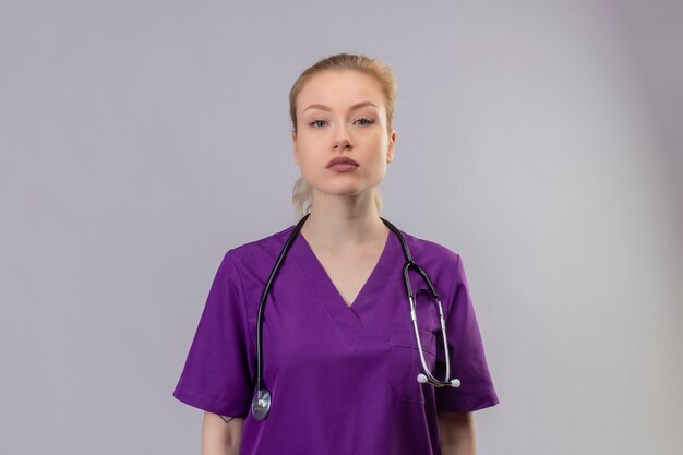 Regardant la caméra jeune médecin portant une robe médicale violette et un stéthoscope sur un mur blanc isolé