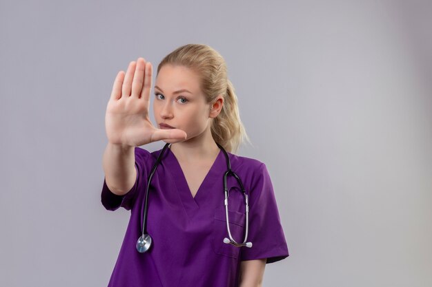 Regardant la caméra jeune médecin vêtu d'une robe médicale violette et stéthoscope montre le geste d'arrêt sur un mur blanc isolé