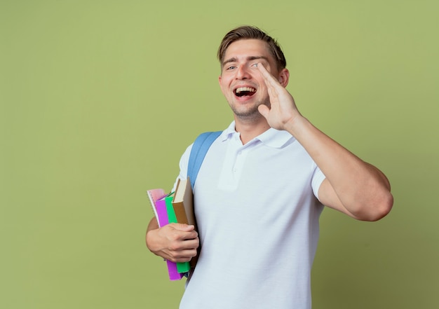 Regardant la caméra joyeux jeune étudiant beau mâle portant un sac à dos tenant des livres et appelant quelqu'un isolé sur fond vert olive