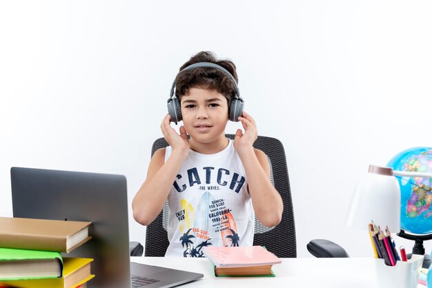 Regardant la caméra petit écolier portant des écouteurs assis au bureau avec des outils scolaires mettant la main sur des écouteurs isolé sur fond blanc