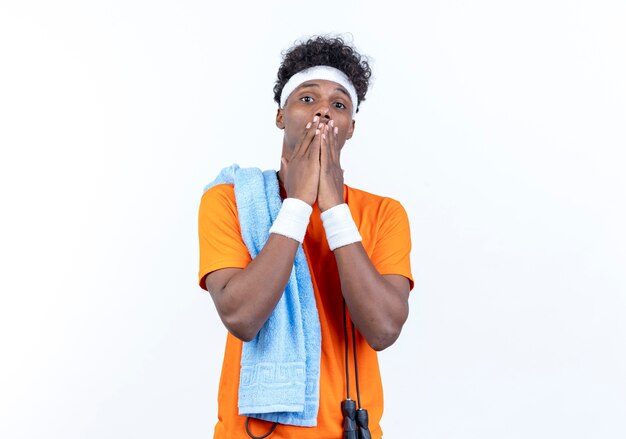 Regardant la caméra peur jeune homme sportif afro-américain portant bandeau et bracelet couvert bouche avec les mains avec une serviette sur l'épaule isolé sur fond blanc