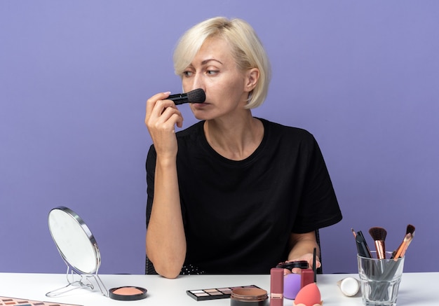 En regardant le côté, une belle jeune fille est assise à table avec des outils de maquillage appliquant un fard à joues en poudre avec un pinceau à poudre isolé sur un mur bleu