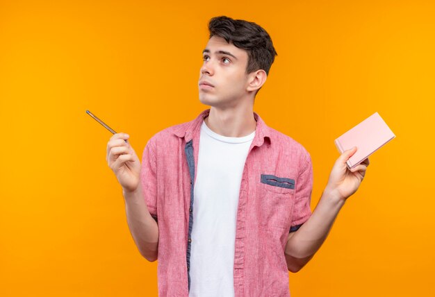 Regardant le côté caucasien jeune homme portant une chemise rose tenant un stylo et un ordinateur portable sur un mur orange isolé