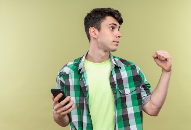 Regardant le côté caucasien jeune homme portant une chemise verte tenant le téléphone montrant oui geste sur mur vert isolé