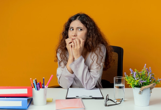 Regardant à côté concerné jeune jolie femme employé de bureau assis au bureau avec des outils de bureau mord les ongles isolés sur le mur orange