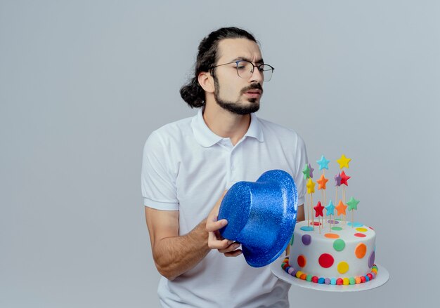 Regardant à côté confus bel homme tenant un gâteau avec un chapeau bleu isolé sur fond blanc