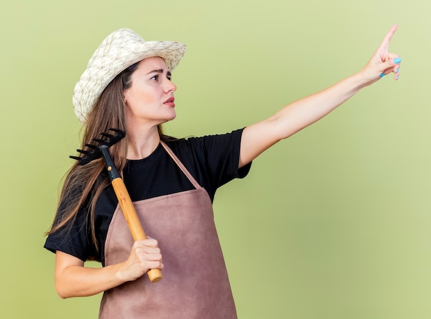 Regardant le côté confus belle fille de jardinier en uniforme portant chapeau de jardinage mettant râteau sur l'épaule et les points sur le côté isolé sur fond vert olive