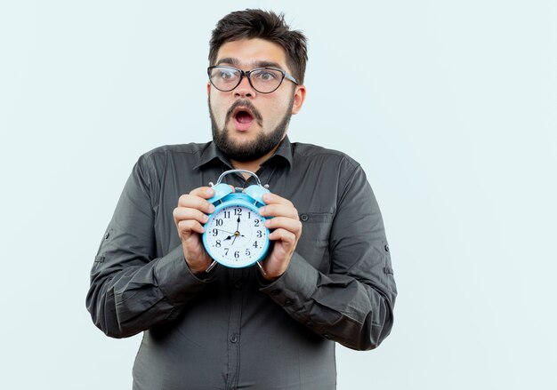 Regardant le côté effrayé jeune homme d'affaires portant des lunettes tenant un réveil isolé sur fond blanc