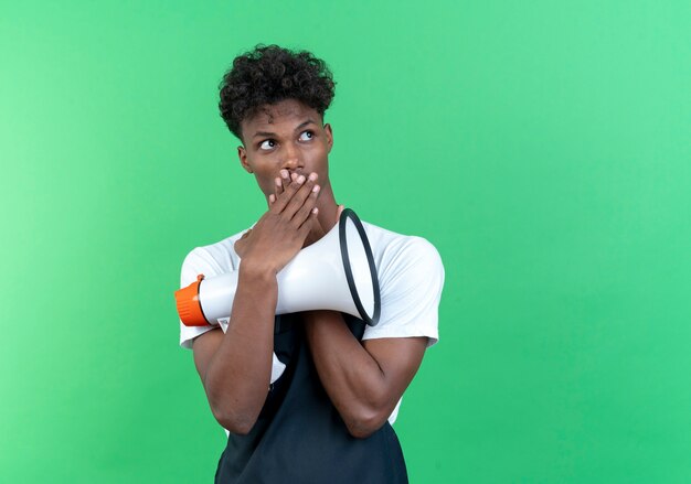 Regardant le côté effrayé jeune homme afro-américain coiffeur portant l'uniforme tenant haut-parleur et bouche couverte avec la main isolé sur fond vert