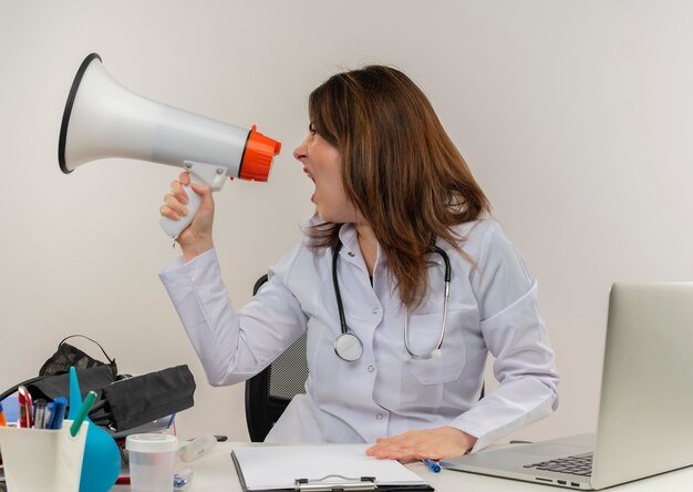 Regardant à côté femme médecin d'âge moyen portant portant une robe médicale avec stéthoscope assis au bureau de travail sur un ordinateur portable avec des outils médicaux parle sur haut-parleur sur mur blanc avec espace de copie