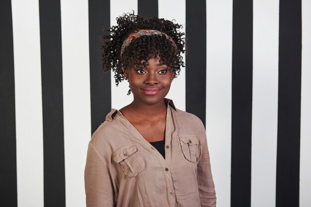 En regardant de côté. Fille afro-américaine souriante se tient dans le studio avec des lignes verticales blanches et noires à l'arrière-plan