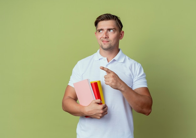 Regardant à côté heureux jeune étudiant beau mâle tenant des livres et des points à côté isolé sur fond vert olive