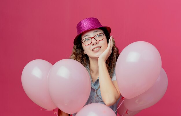 Regardant à côté heureux jeune fille portant des lunettes et un chapeau rose debout derrière des ballons et mettant la main sur la joue isolé sur fond rose