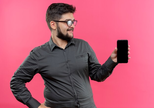 Regardant à côté heureux jeune homme d'affaires portant des lunettes tenant le téléphone à côté isolé sur mur rose