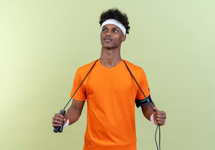 Photo gratuit regardant à côté heureux jeune homme sportif afro-américain portant bandeau et bracelet avec corde à sauter sur l'épaule isolé sur fond vert