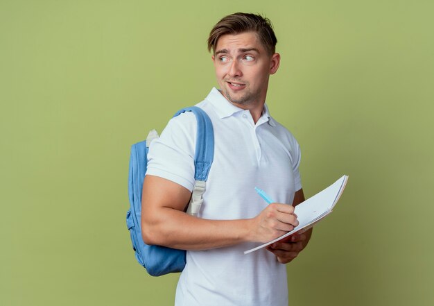 Regardant à côté impressionné jeune beau étudiant masculin portant un sac à dos tenant un ordinateur portable avec un stylo
