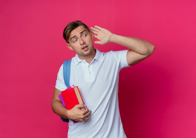 Regardant le côté jeune beau étudiant masculin portant un sac à dos tenant des livres et montrant le geste d'écoute isolé sur un mur rose