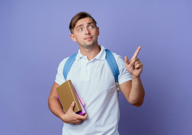 Regardant le côté jeune beau étudiant masculin portant un sac à dos tenant des livres et des points sur le côté isolé sur un mur bleu avec espace copie