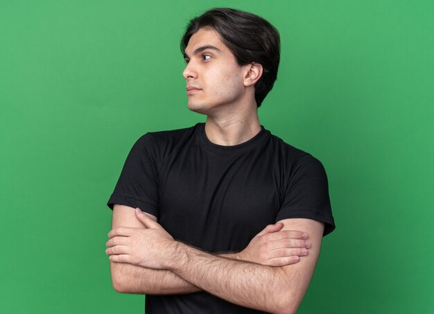 Regardant le côté jeune beau mec portant un t-shirt noir croisant les mains isolés sur le mur vert