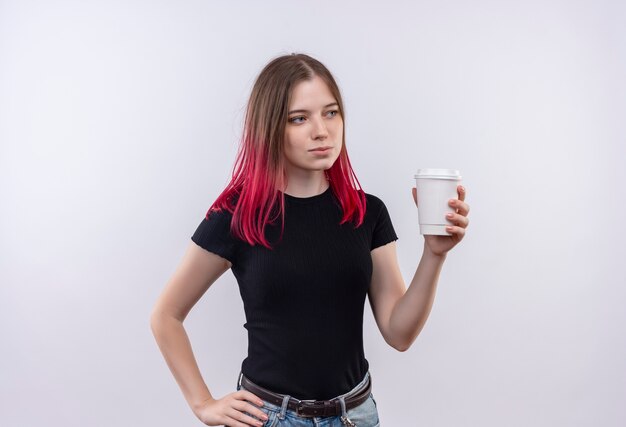 Regardant le côté jeune belle fille portant un t-shirt noir tenant une tasse de café mettant sa main sur la hanche sur fond blanc isolé