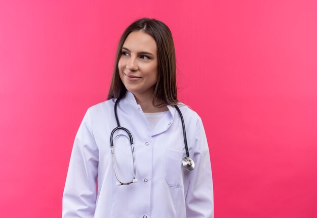 Regardant à côté jeune fille médecin portant robe médicale stéthoscope sur fond rose isolé