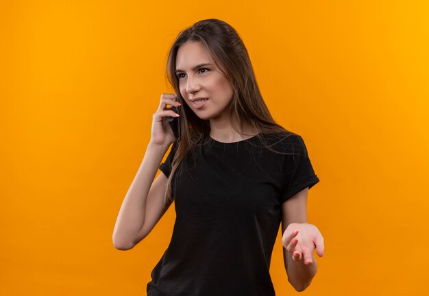 Regardant à côté jeune fille de race blanche portant un t-shirt noir parle au téléphone sur fond orange isolé