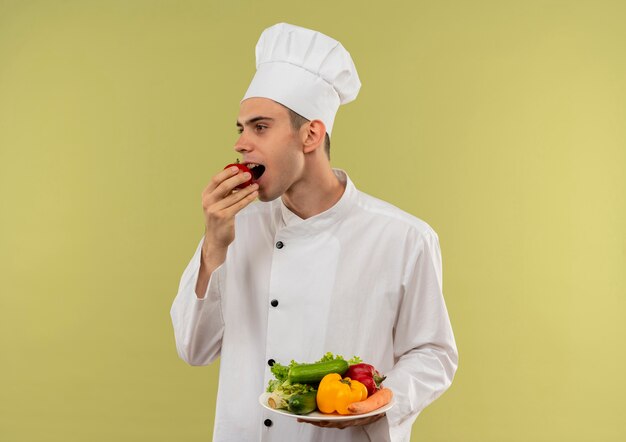 Regardant à côté jeune homme cuisinier portant un uniforme de chef tenant des légumes sur la plaque mord la tomate sur un mur vert isolé avec copie espace