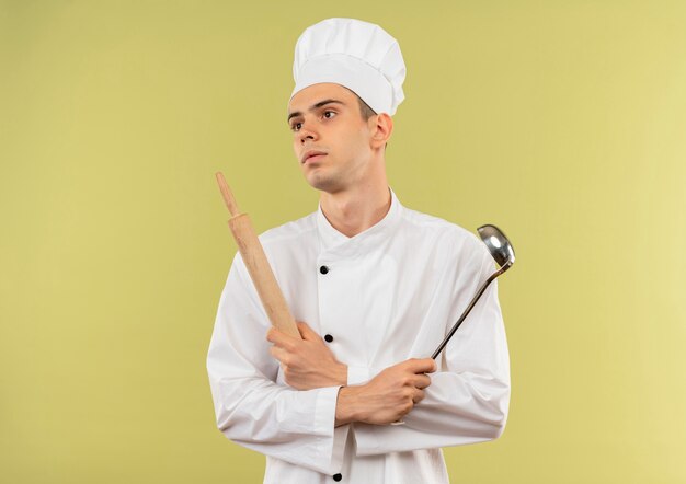 Regardant à côté jeune homme cuisinier portant l'uniforme du chef traversant la louche et le rouleau à pâtisserie dans sa main sur un mur vert isolé avec copie espace