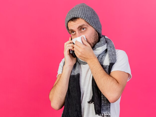 Regardant à côté jeune homme malade portant chapeau d'hiver avec écharpe parle au téléphone et essuyant le nez avec serviette isolé sur fond rose avec espace copie