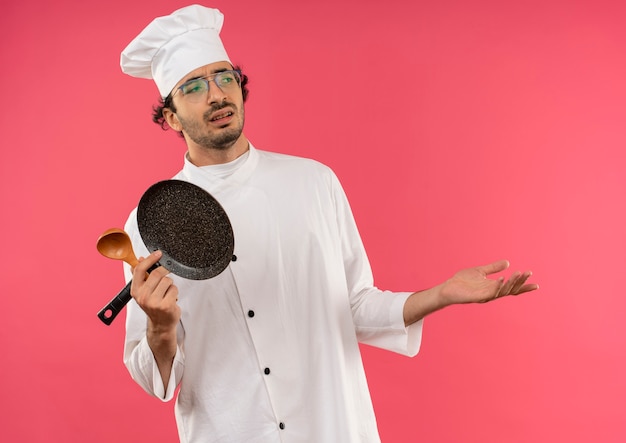 Photo gratuite regardant à côté un jeune homme mécontent cuisinier portant l'uniforme de chef et des verres tenant une poêle avec une cuillère et une main répandue