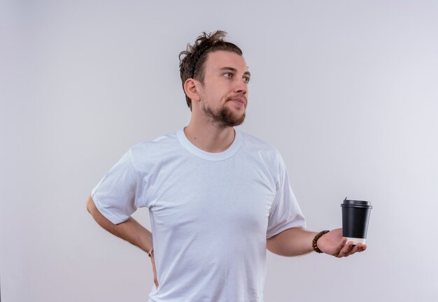 Regardant à côté jeune mec portant un t-shirt blanc tenant une tasse de café a mis sa main sur la hanche sur fond blanc isolé