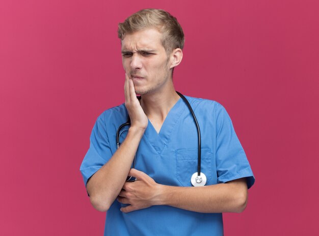 En regardant de côté un jeune médecin de sexe masculin portant un uniforme de médecin avec un stéthoscope mettant la main sur une dent douloureuse isolée sur un mur rose