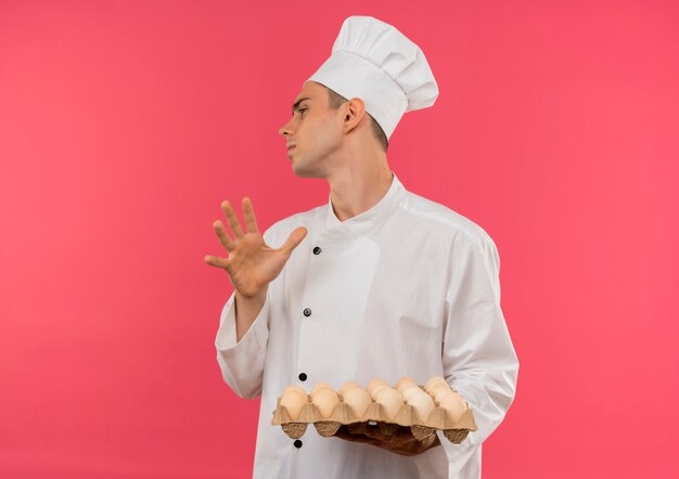 Regardant à côté les jeunes hommes cuisinier portant l'uniforme de chef holding lot d'oeufs montrant le geste d'arrêt sur mur rose isolé