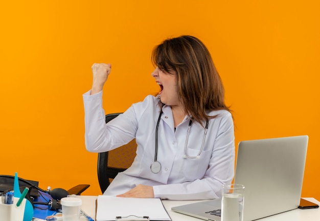 Regardant à côté joyeuse femme médecin d'âge moyen portant une robe médicale avec stéthoscope assis au bureau de travail sur un ordinateur portable avec des outils médicaux faisant un geste fort sur un mur orange avec espace de copie