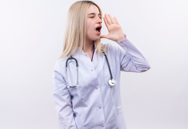 Regardant à côté médecin jeune fille blonde portant un stéthoscope et une robe médicale appelant quelqu'un sur un mur blanc isolé