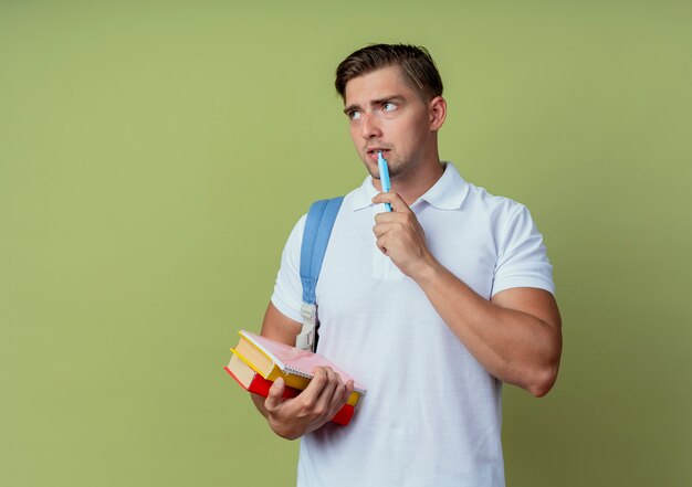 Regardant le côté pensant jeune beau étudiant masculin portant un sac à dos tenant des livres et mettant un stylo sur la bouche isolé sur fond vert olive