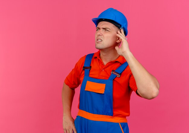 Regardant le côté pensant jeune constructeur de sexe masculin portant l'uniforme et un casque de sécurité mettant la main sur l'oreille