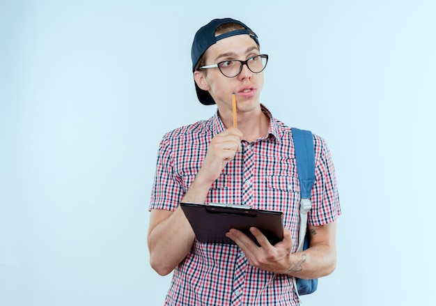 Regardant le côté pensant jeune garçon étudiant portant sac à dos et lunettes et casquette tenant le presse-papiers et en gardant le stylo sur la joue