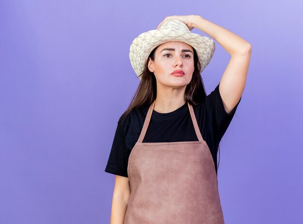 Regardant côté pensée belle fille de jardinier en uniforme portant chapeau de jardinage mettant la main sur le chapeau isolé sur fond bleu avec espace de copie