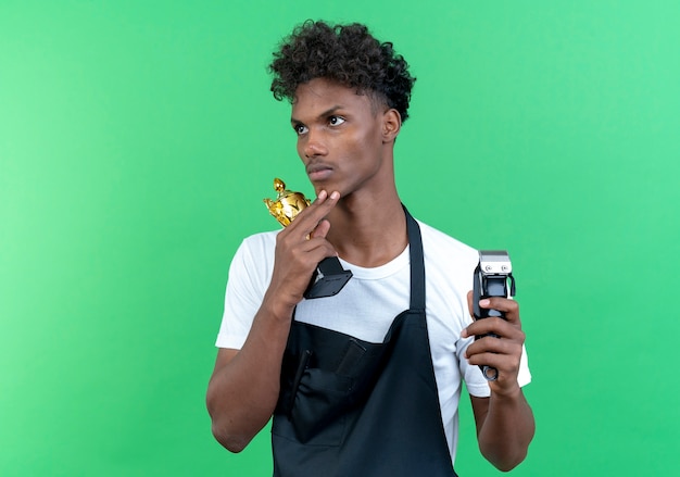 Regardant à côté de la pensée jeune barbier afro-américain portant l'uniforme tenant la coupe du gagnant avec des tondeuses à cheveux isolé sur fond vert