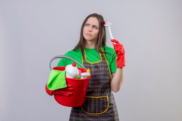 Regardant côté pensée nettoyage jeune femme portant l'uniforme dans des gants rouges tenant des outils de nettoyage rayures tête avec brosse de nettoyage sur mur blanc isolé