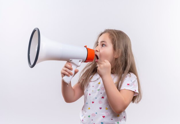 Regardant à côté petite écolière portant un t-shirt blanc parle haut-parleur creux sur mur blanc isolé