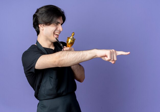 Regardant le côté souriant jeune beau coiffeur masculin en uniforme tenant la coupe du gagnant et les points à côté isolé sur le mur bleu