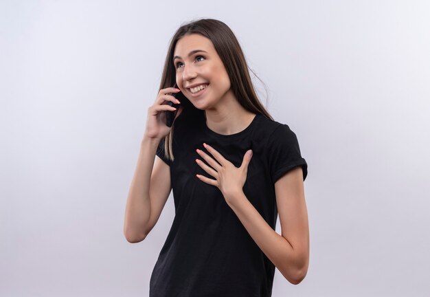 Regardant à côté souriant jeune fille de race blanche portant un t-shirt noir speakes sur téléphone met sa main sur le coeur sur un mur blanc isolé