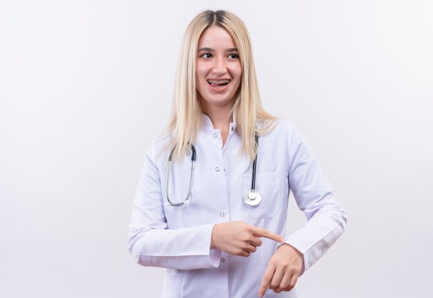 Regardant à côté souriant médecin jeune fille blonde portant un stéthoscope et une robe médicale en orthèse dentaire montrant le geste de montre-bracelet sur mur blanc isolé