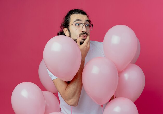 Regardant à côté surpris bel homme portant des lunettes debout parmi les ballons et mettant la main sur la joue isolé sur fond rose