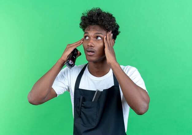 En regardant le côté surpris, un jeune coiffeur afro-américain portant un uniforme tenant une tondeuse à cheveux et mettant les mains sur le front