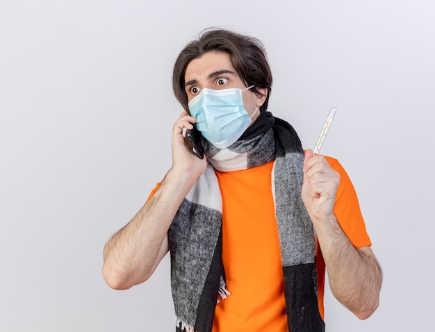 Regardant à côté surpris jeune homme malade portant un foulard et un masque médical parle au téléphone et tenant un thermomètre isolé sur fond blanc