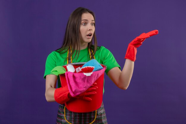 Regardant à côté surpris nettoyage jeune femme en uniforme dans des gants rouges tenant des outils de nettoyage levant la main sur un mur violet isolé